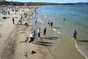 Plaża pełna ludzi, niektórzy wchodzą do wody, inni spacerują po brzegu. Słoneczna pogoda.