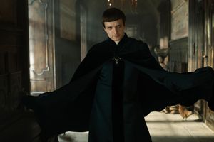 Mężczyzna w czarnym pelerynie, z zamkniętymi drzwiami w tle, wygląda tajemniczo.