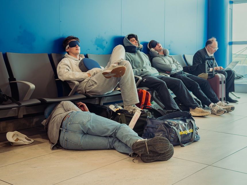 Grupa osób w wygodnych ubraniach śpi w poczekalni lotniska na plastikowych ławkach.