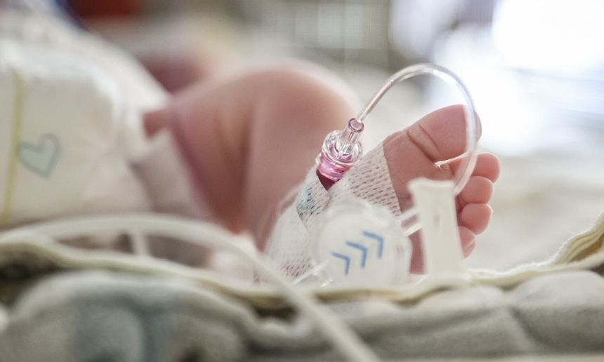 Zdjęcie przedstawia stopę noworodka z rurkami medycznymi w otoczeniu szpitalnym.