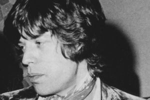 Mężczyzna z długimi włosami, w eleganckim ubiorze, z chustą na szyi, patrzy w bok.
