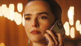 Kobieta trzymająca telefon, z zamyślonym spojrzeniem, w tle rozmyte światła.