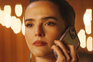 Kobieta trzymająca telefon, z zamyślonym spojrzeniem, w tle rozmyte światła.