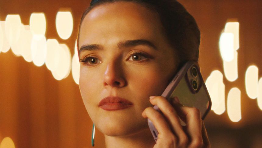 Kobieta trzymająca telefon, z zamyślonym spojrzeniem, w tle rozmyte światła.