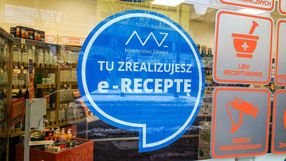 Na zdjęciu widoczny jest niebieski znaczek z napisem „TU ZREALIZUJESZ e-RECEPTĘ”. W tle apteka.