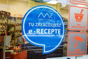 Na zdjęciu widoczny jest niebieski znaczek z napisem „TU ZREALIZUJESZ e-RECEPTĘ”. W tle apteka.