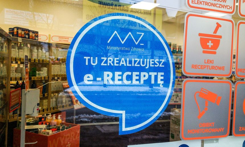 Na zdjęciu widoczny jest niebieski znaczek z napisem „TU ZREALIZUJESZ e-RECEPTĘ”. W tle apteka.