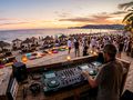 DJ gra na plaży podczas zachodu słońca, ludzie tańczą, relaksują się na leżakach.