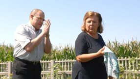 Dwoje ludzi w polu, mężczyzna złożonymi dłońmi, kobieta z szarfą w ręku. Tło: kukurydza.