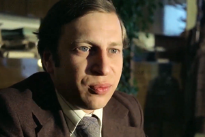 Mężczyzna w eleganckim ubraniu, siedzący na tle półek z książkami, patrzy w obiektyw.