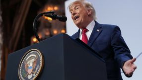 Mężczyzna w garniturze mówi do mikrofonu, na tle podium z godłem USA.