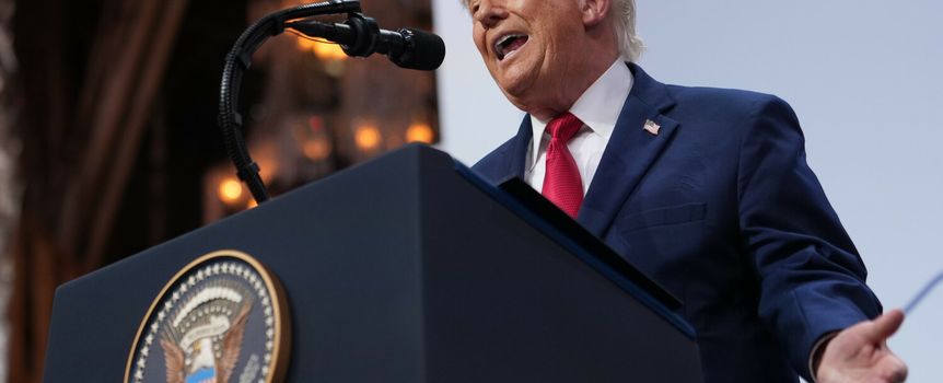 Mężczyzna w garniturze mówi do mikrofonu, na tle podium z godłem USA.