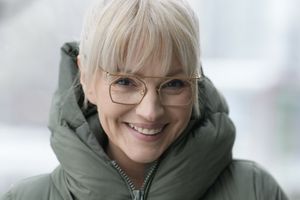 Uśmiechnięta kobieta w dużej kurtce zimowej i okularach, w szarej scenerii zimowej.