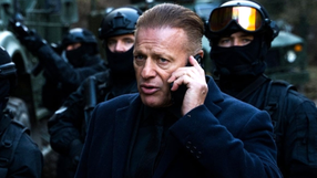 Mężczyzna w eleganckim ubraniu, rozmawia przez telefon, w tle zamaskowani ochroniarze.