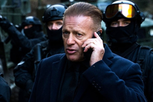 Mężczyzna w eleganckim ubraniu, rozmawia przez telefon, w tle zamaskowani ochroniarze.