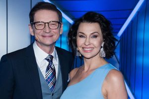 Na zdjęciu uśmiechnięta para w eleganckich ubraniach na tle niebieskiego oświetlenia.
