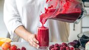 Osoba w białej bluzie nalewa czerwony smoothie do szklanki, obok owoce na stole.