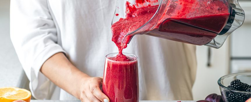 Osoba w białej bluzie nalewa czerwony smoothie do szklanki, obok owoce na stole.