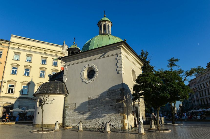 Widok na mały budynek z zieloną kopułą, otoczony drzewami i ludźmi w słoneczny dzień.