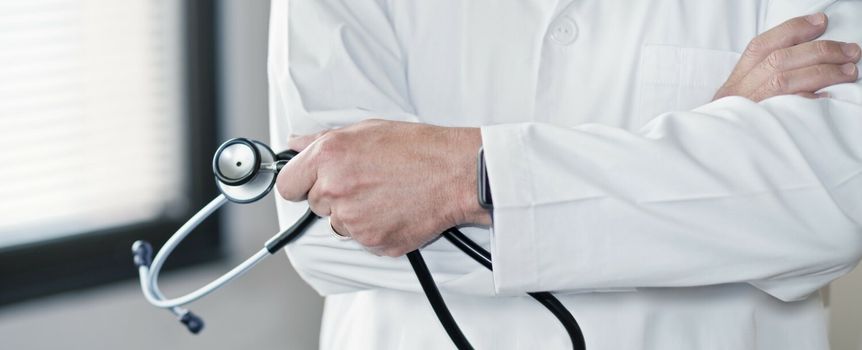 Mężczyzna w białym fartuchu trzyma stetoskop, z założonymi rękami w dłoniach.