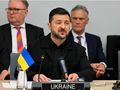 Prezydent Ukrainy Wołodymyr Zełenski przemawia, siedząc przy stole, z flagą Ukrainy obok.