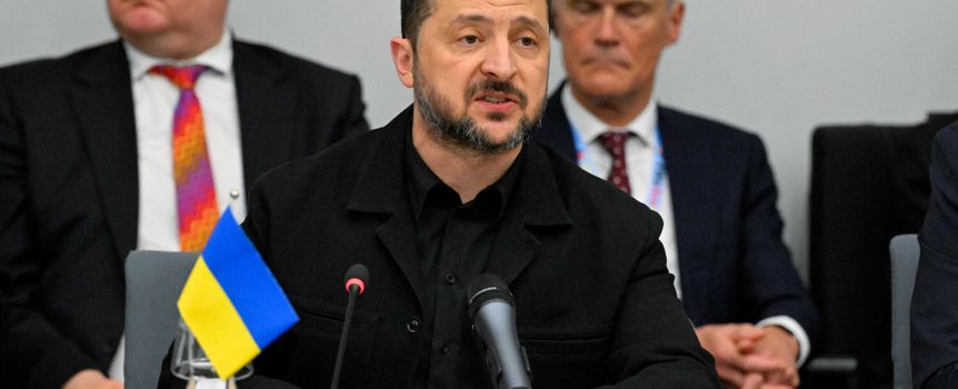 Prezydent Ukrainy Wołodymyr Zełenski przemawia, siedząc przy stole, z flagą Ukrainy obok.