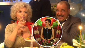 Na zdjęciu dwie osoby przy stole, jedna pije z kieliszka, obok uśmiechnięty mężczyzna, mikrofon w tle.