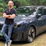 Mężczyzna w czarnej koszulce stoi obok granatowego SUV-a w otoczeniu drzew.