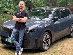 Mężczyzna w czarnej koszulce stoi obok granatowego SUV-a w otoczeniu drzew.