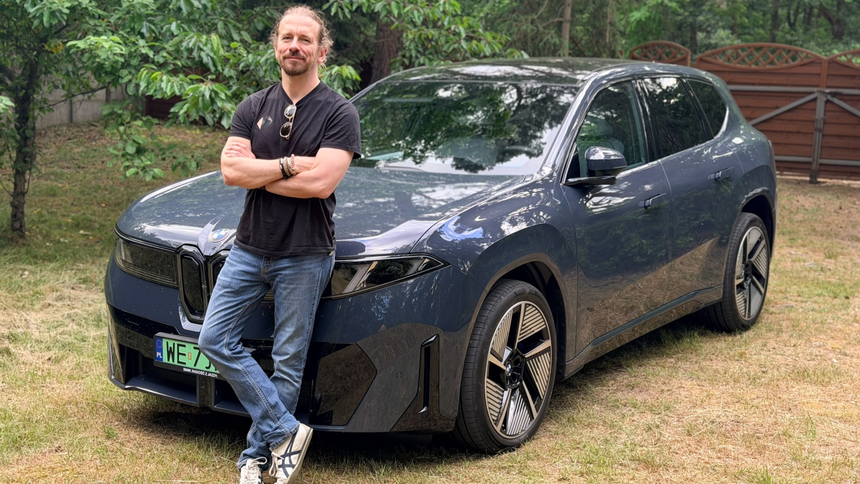 Mężczyzna w czarnej koszulce stoi obok granatowego SUV-a w otoczeniu drzew.
