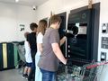 Na zdjęciu dzieci oddają opakowania do maszyny recyklingowej w punkcie zbiórki.