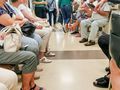 Zdjęcie przedstawia poczekalnię z siedzącymi ludźmi, w różnych ubraniach, rozmawiającymi ze sobą.