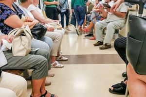Zdjęcie przedstawia poczekalnię z siedzącymi ludźmi, w różnych ubraniach, rozmawiającymi ze sobą.