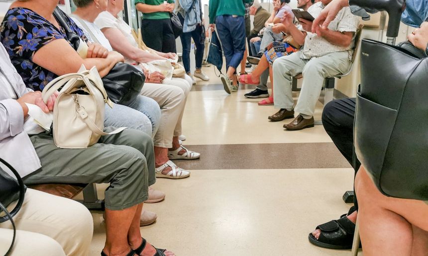 Zdjęcie przedstawia poczekalnię z siedzącymi ludźmi, w różnych ubraniach, rozmawiającymi ze sobą.