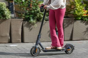 Osoba na hulajnodze elektrycznej, ubrana w jasny sweter i różowe legginsy, na tle roślinności.