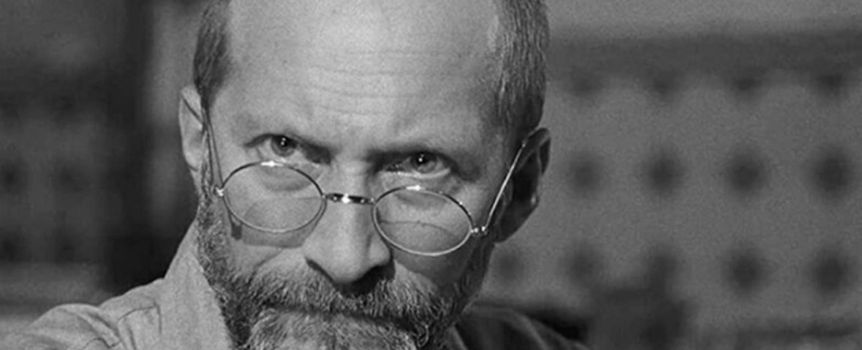 Mężczyzna z brodą i okularami, patrzy intensywnie, w tle rozmyte wzory. Czarno-biała fotografia.