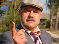 Mężczyzna w kapeluszu, z wąsem, wskazuje palcem, ma groźny wyraz twarzy. Tło to zieleń i budynki.