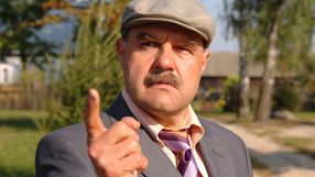 Mężczyzna w kapeluszu, z wąsem, wskazuje palcem, ma groźny wyraz twarzy. Tło to zieleń i budynki.
