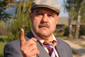 Mężczyzna w kapeluszu, z wąsem, wskazuje palcem, ma groźny wyraz twarzy. Tło to zieleń i budynki.