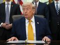 Na zdjęciu Donald Trump w garniturze, siedzący przy biurku z dokumentami. W tle inni mężczyźni.