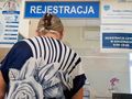 Kobieta w kwiecistej bluzce stoi przy rejestracji w placówce medycznej. Na ścianie widoczne napisy.