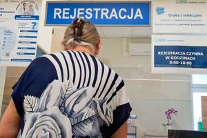 Kobieta w kwiecistej bluzce stoi przy rejestracji w placówce medycznej. Na ścianie widoczne napisy.
