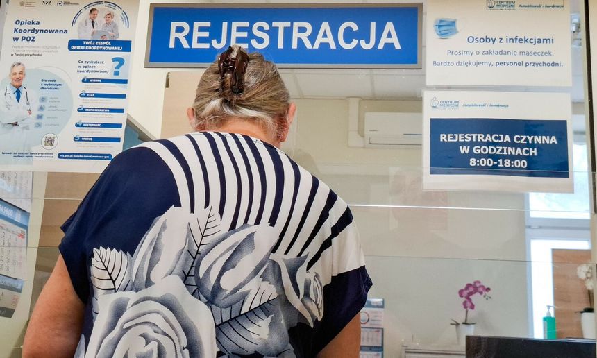Kobieta w kwiecistej bluzce stoi przy rejestracji w placówce medycznej. Na ścianie widoczne napisy.