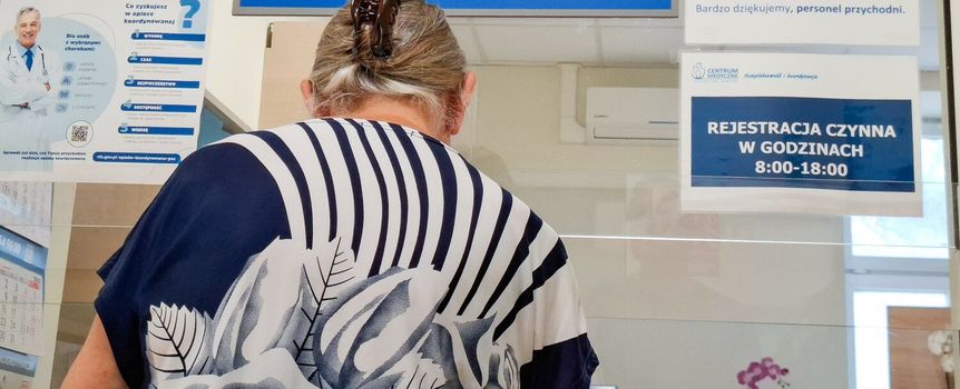 Kobieta w kwiecistej bluzce stoi przy rejestracji w placówce medycznej. Na ścianie widoczne napisy.