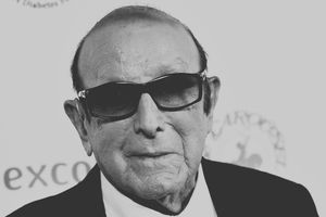 Mężczyzna w ciemnych okularach, uśmiechnięty, w eleganckim garniturze. Tło białe z napisami.