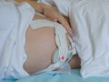Na zdjęciu widać ciężarną kobietę leżącą na łóżku, z ręką na brzuchu i założonym aparatem monitorującym.