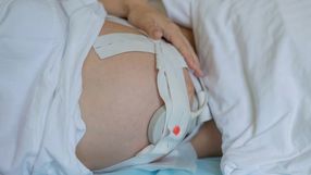 Na zdjęciu widać ciężarną kobietę leżącą na łóżku, z ręką na brzuchu i założonym aparatem monitorującym.