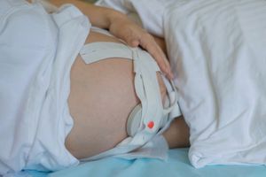 Na zdjęciu widać ciężarną kobietę leżącą na łóżku, z ręką na brzuchu i założonym aparatem monitorującym.