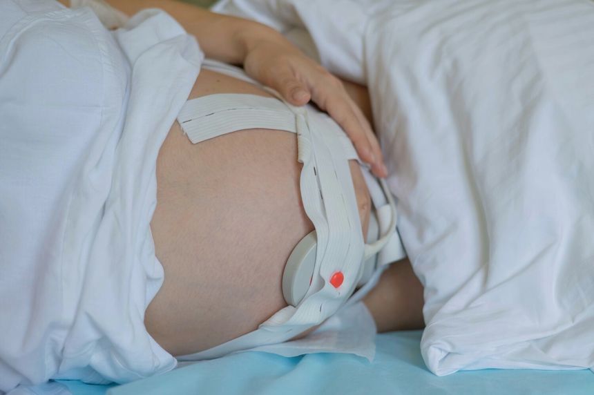 Na zdjęciu widać ciężarną kobietę leżącą na łóżku, z ręką na brzuchu i założonym aparatem monitorującym.