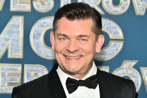 Na zdjęciu mężczyzna w czarnym garniturze z muszką, uśmiechnięty, na tle niebieskiego napisu.